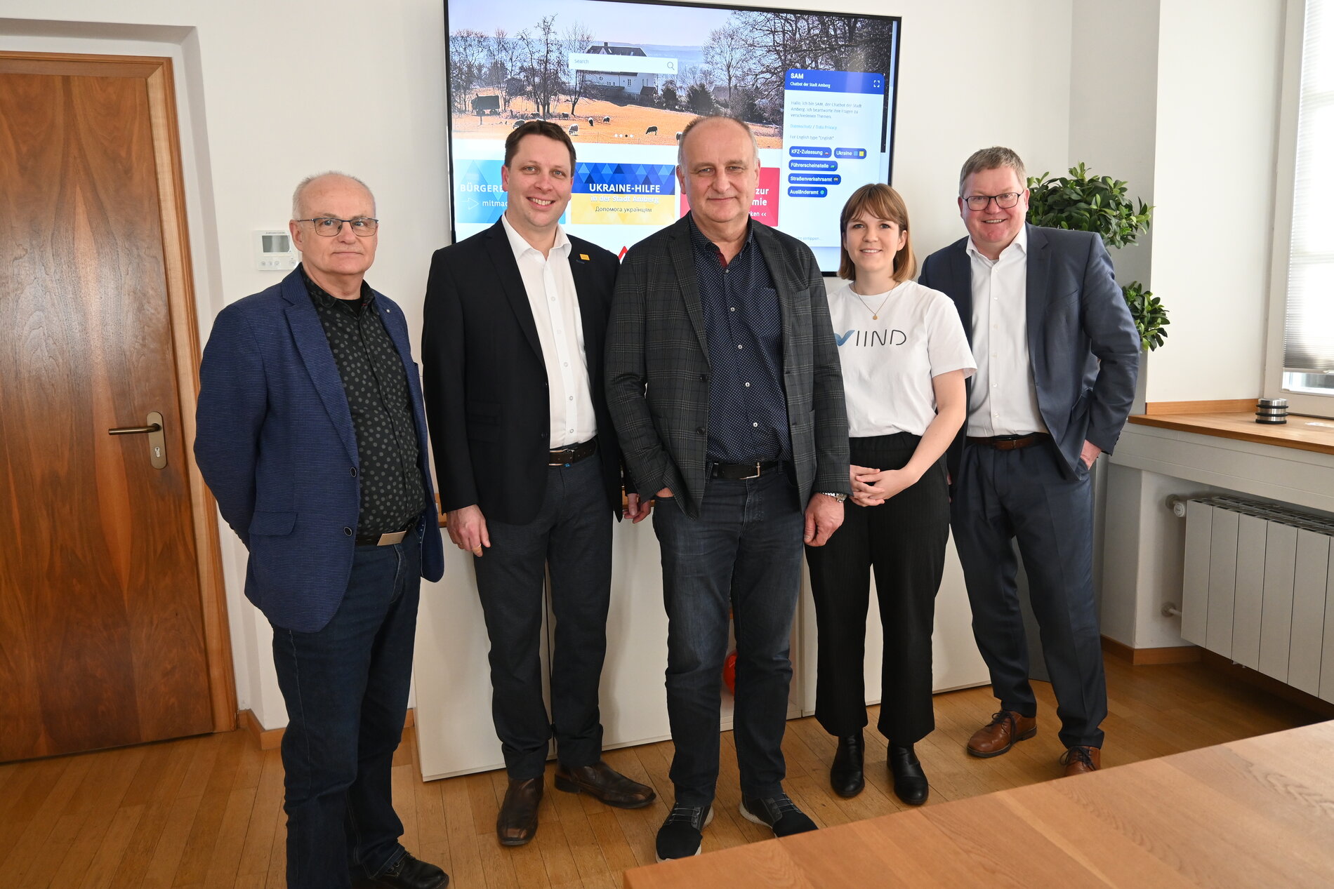 Vorstellung von SAM, dem Chatbot der Stadt Amberg, durch Josef Singer (Leiter der Stabsstelle Zentrale Dienste), Martin Schafbauer (Leiter Einwohneramt), Markus Willecke (IT-Abteilung), Marie Hartz (viind GmbH), Oberbürgermeister Michael Cerny und im Hintergrund SAM. Foto © Thomas Graml, Stadt Amberg